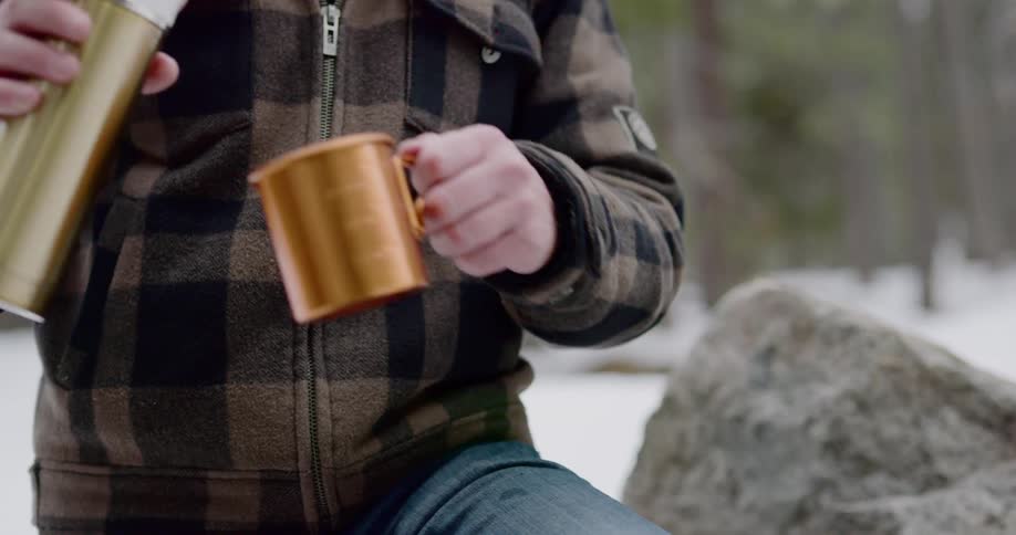 Man sitting on a big rock pours tea out of a thermos bottle in the snow covered woods