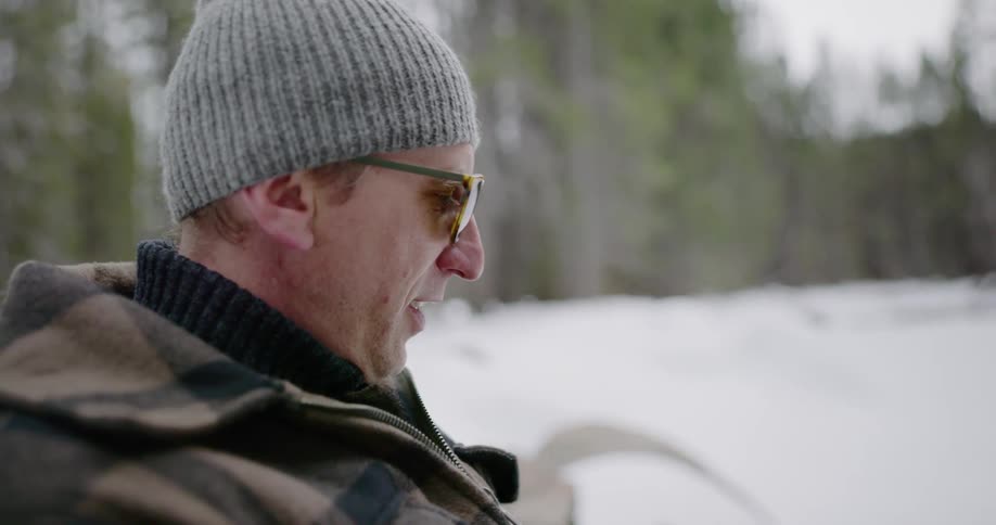A man with sunglasses and a hat drinking tea with snow and trees in the background