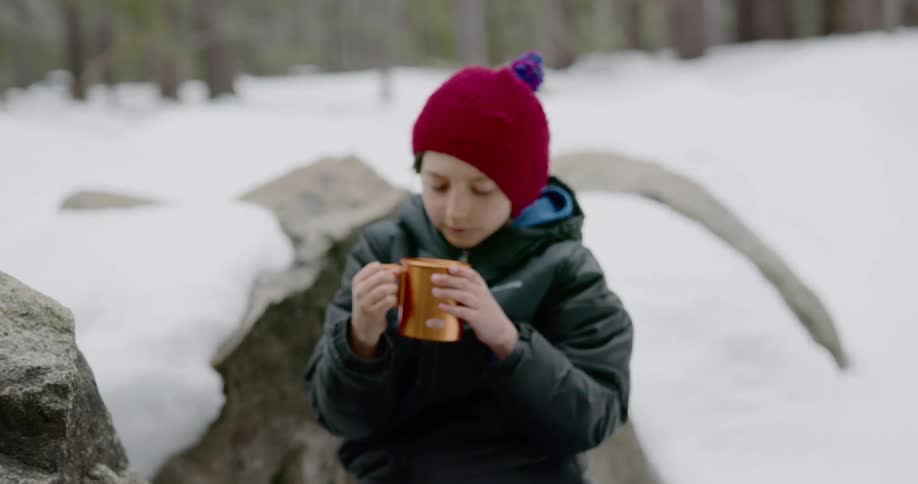 Father and son sit on a rock and pour tea out of thermos bottle in the snow covered woods