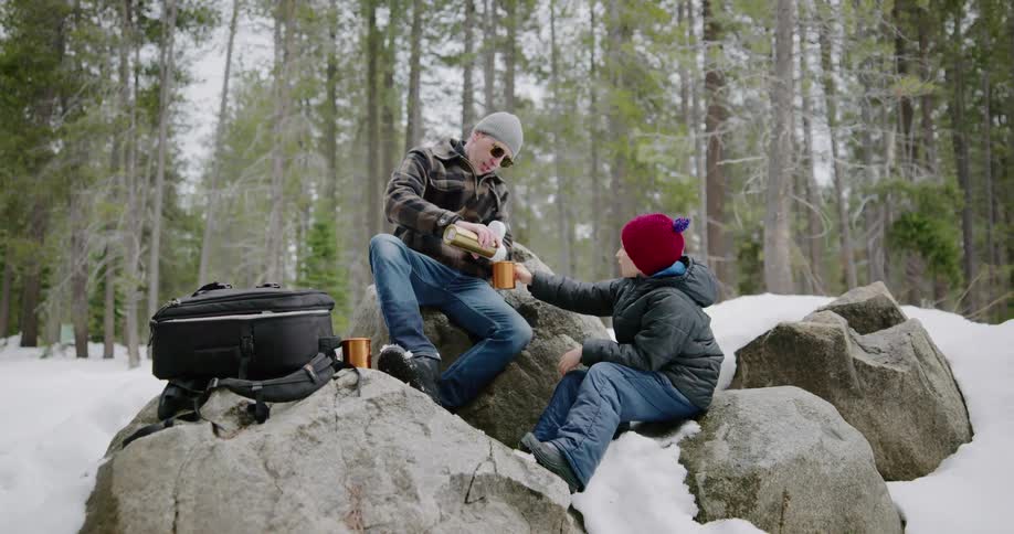 Man and boy sit on large stones among snow covered clearing near forest