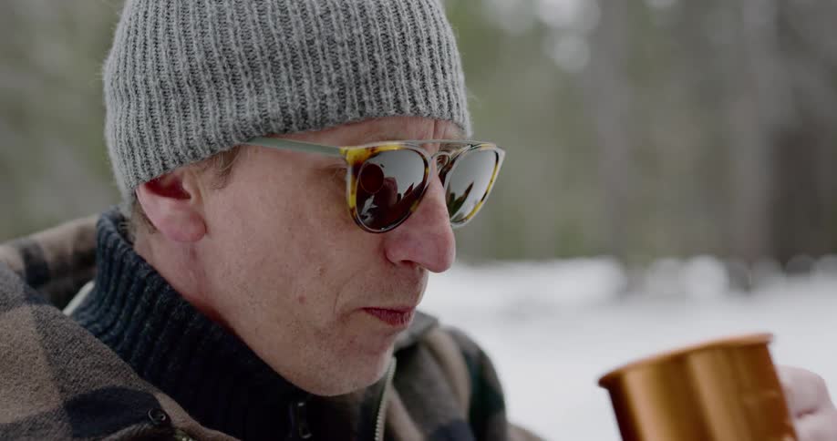 A man drinks from a mug while outside in warm clothes in the snow