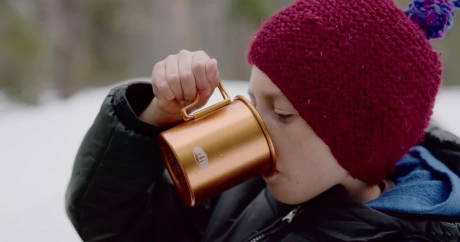 Boy in a red beanie drinking from copper mug outdoors in the snow