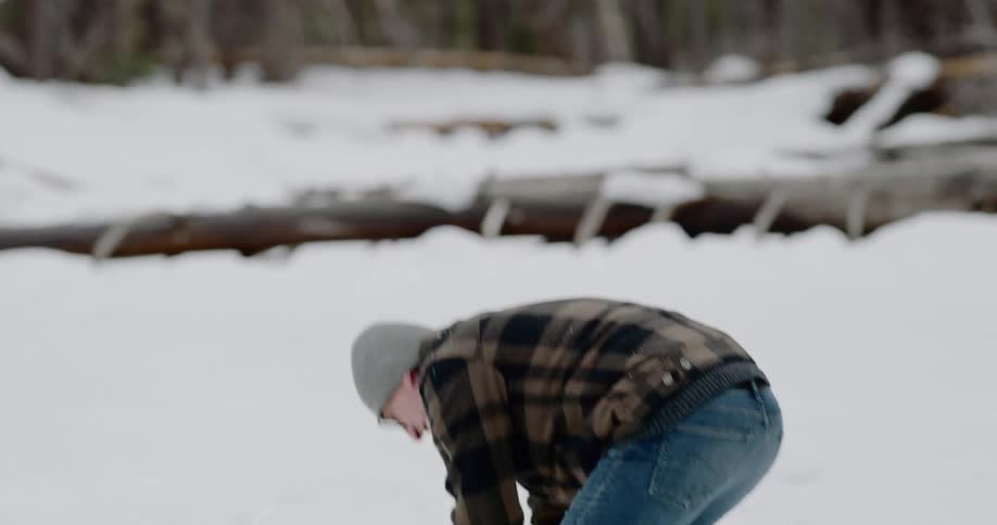 Male wearing sunglasses bends over to pack snow together then throws the snowball while laughing and shaking off his hands