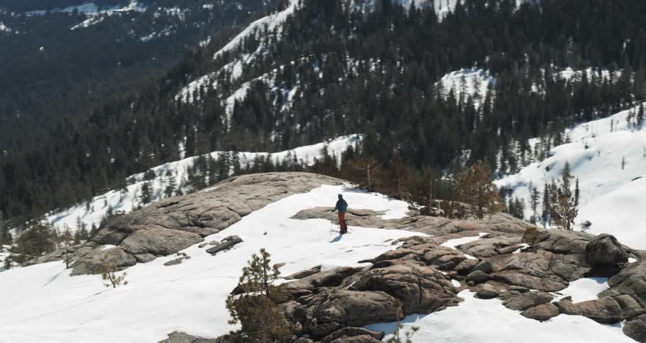 A person stands on top of a large snow covered mountain looking towards a lake and forest