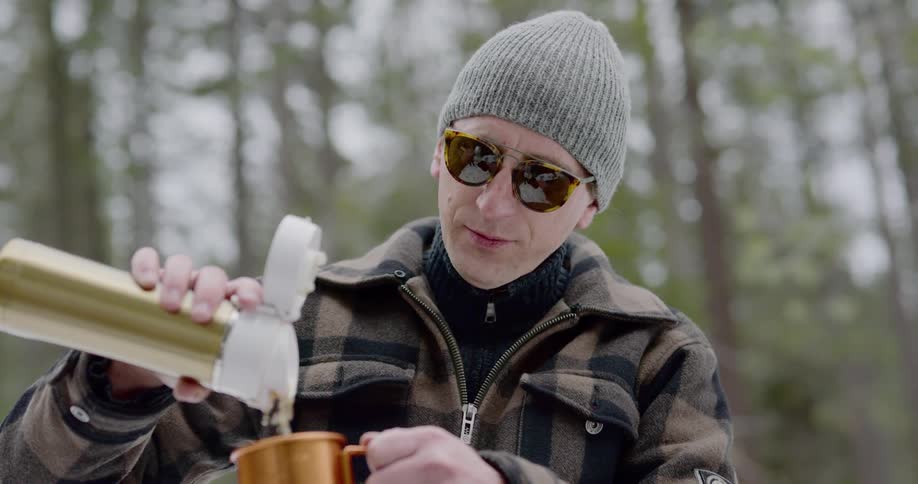 Man sitting on a rock pours hot tea out of thermos bottle in the snow covered woods