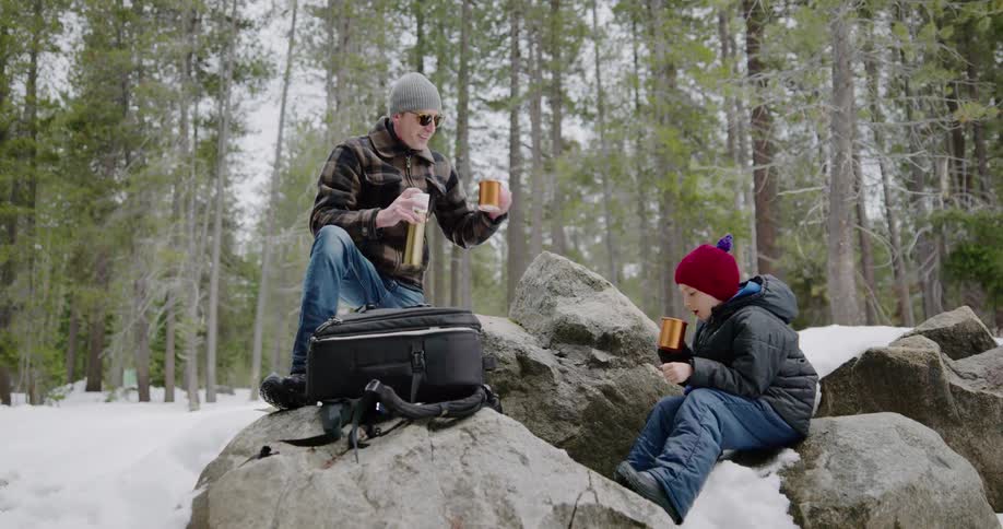 A man and child cheers drinking mugs outside the the snow covered woods