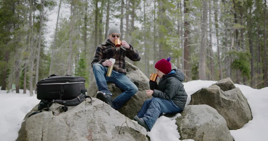 Father and son sit on rocks and pour hot drink out of thermos bottle in the snow covered woods
