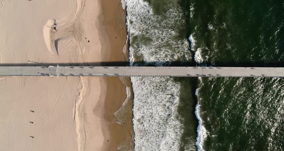 Aerial view of the ocean waves crashing against the beach and a pier sticking out from the land over the sea