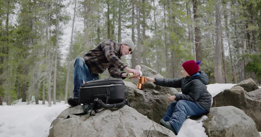Father and son sit on rocks and pour hot drink out of thermos bottle in the snow covered woods