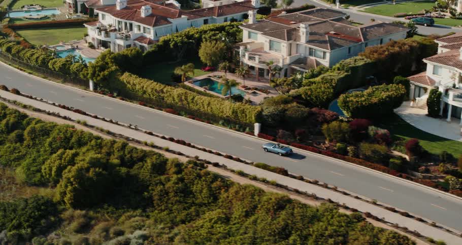 A car drives down a street behind large houses with swimming pools