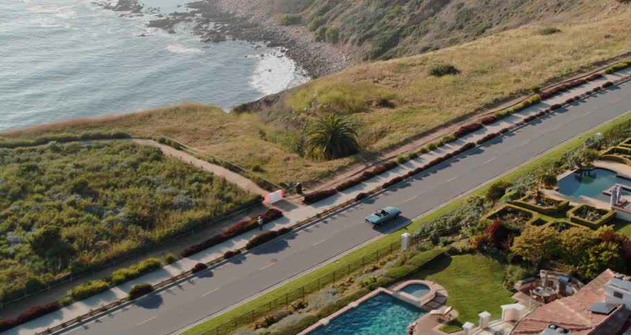 A blue convertible drives down a road next to a cliffside neighborhood that overlooks the ocean