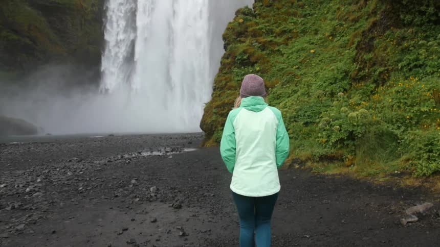 A woman is walking over rocks towards a waterfall