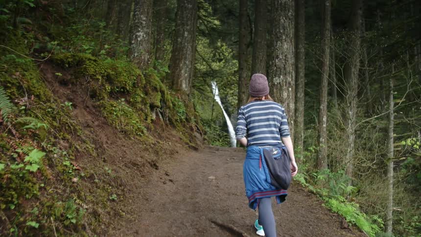 A woman hikes a forest trail towards a waterfall