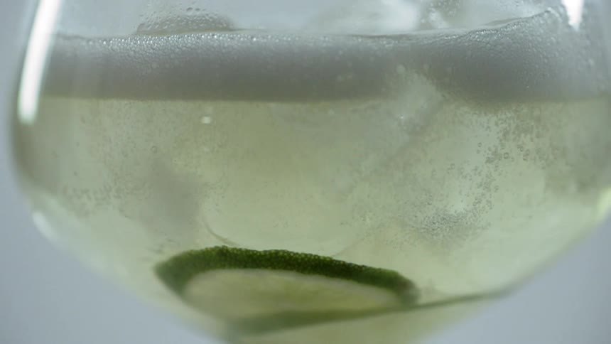 Slices of lime sit at the bottom of a glass with ice as it is filled with sparkling liquid and bubbles rise