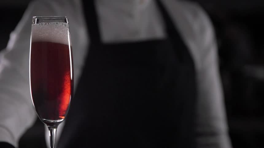 A champagne glass with a red liquid with a person with a white shirt and a black apron in the background