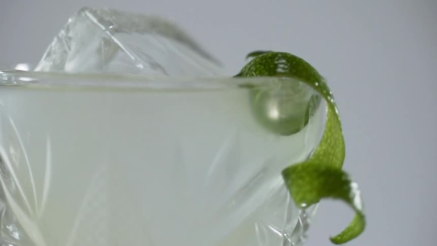 A lime peel falls onto the rim of a glass with ice and alcohol