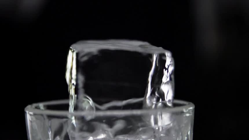 An ice cube hits the side of a clear glass and twirls and spins