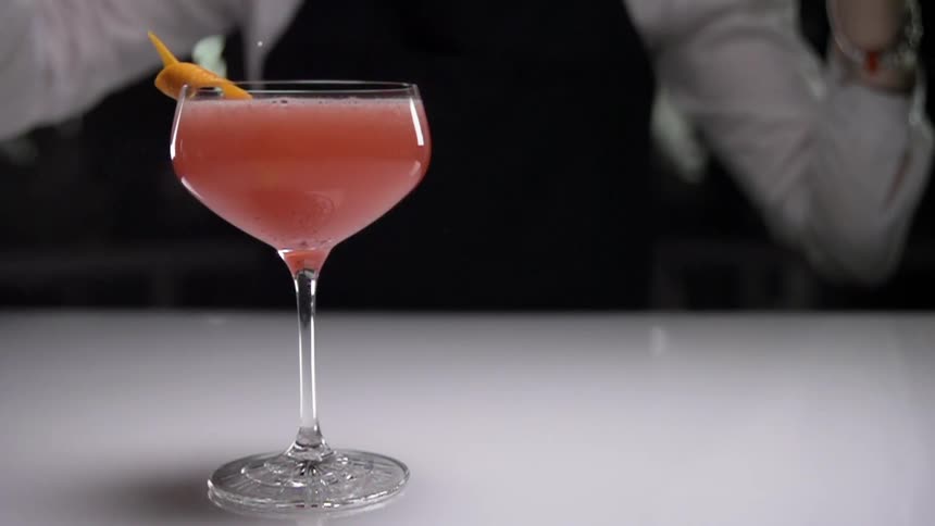 An orange peel is added to a cocktail glass as a bartender stands nearby with her arms up