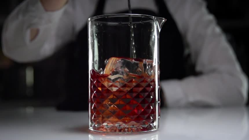 A bartender wearing and apron and white shirt stirs a drink in a mixing glass with a long handled spoon