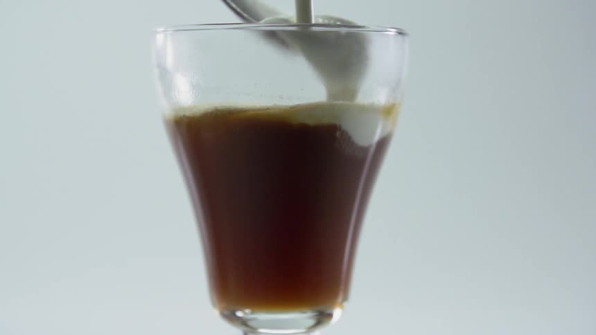Cream is poured over a spoon and into a glass filled with brown liquid