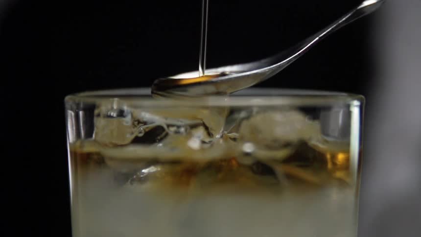 An amber liquid pours over a spoon and falls into a glass filled with ice cubes