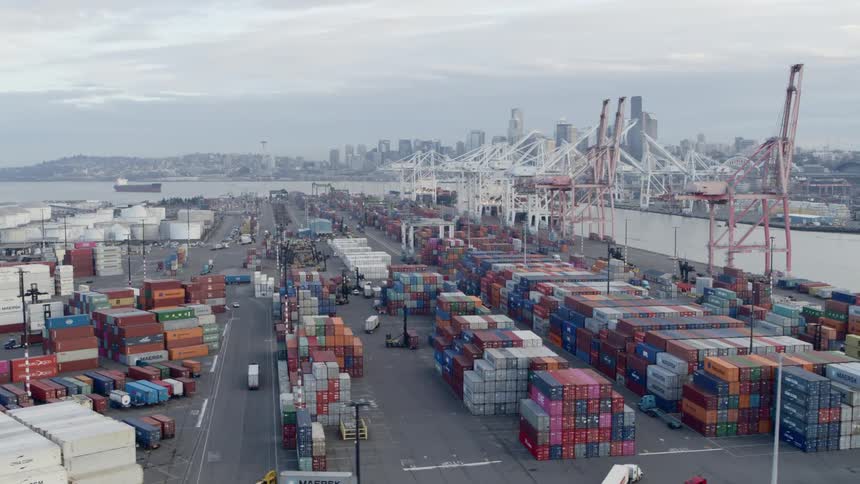 Trucks and heavy equipment drive through stacks of shipping containers at a cargo terminal