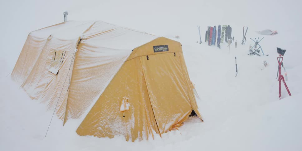 Snow falls on a large yellow multi person tent with skis lined up beside it
