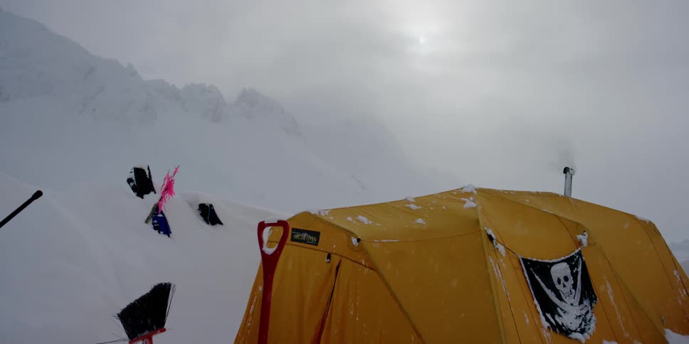 A tent and assorted gear sits near a tall range of snow covered mountains
