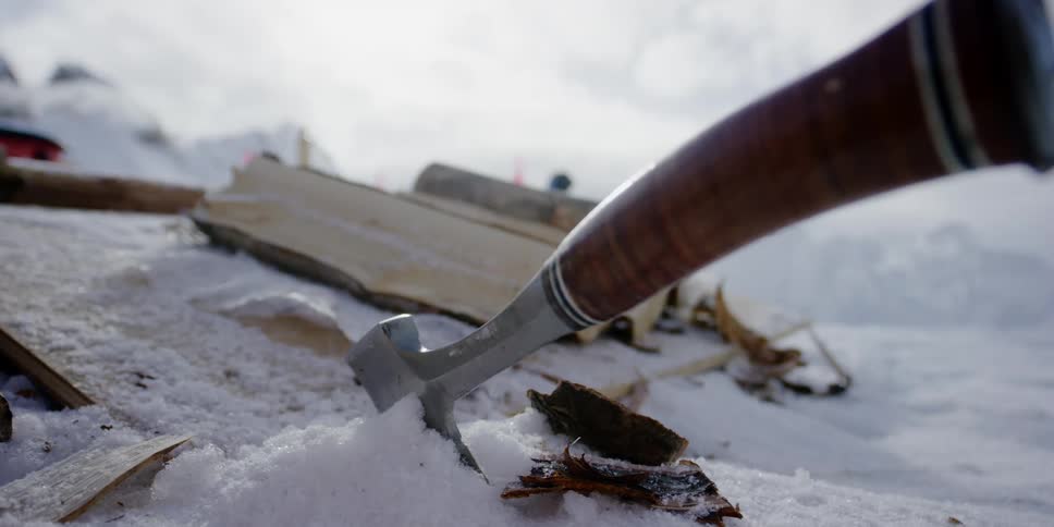 An ax with a wooden handle sticking out of the snow with smoke from a chimney billowing beyond