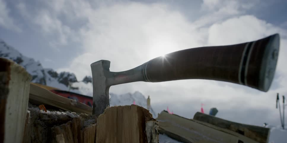 An ax with a wooden handle stuck in wood with smoke billowing from a cabin's chimney