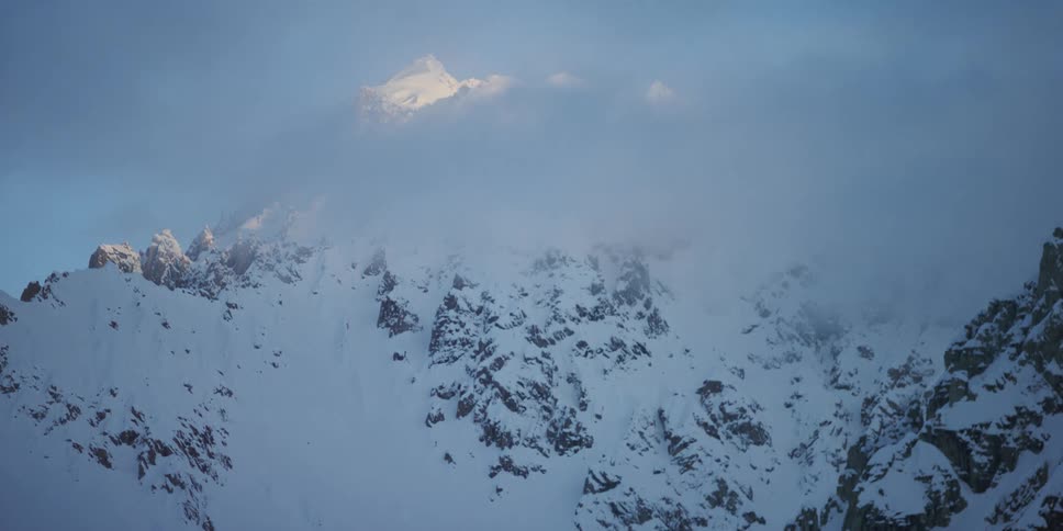 A cloud obscures the snow covered rocky mountain peak on a blue sky sunny day