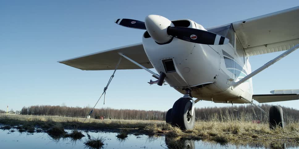 A plane parked near a body of water as another plane takes off in the background