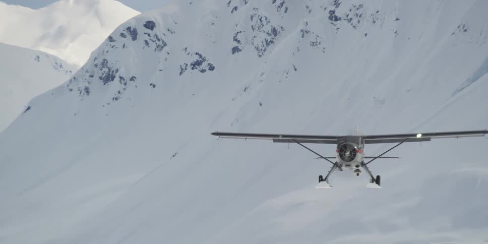 A propeller plane flies over a snow covered mountain slope on a sunny day