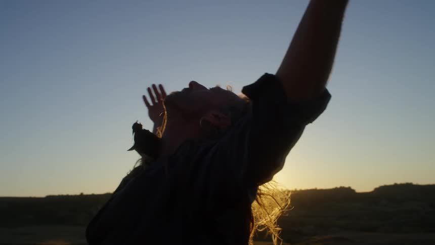 A white man stands in a grassland and holds his hands and arms up to the sky