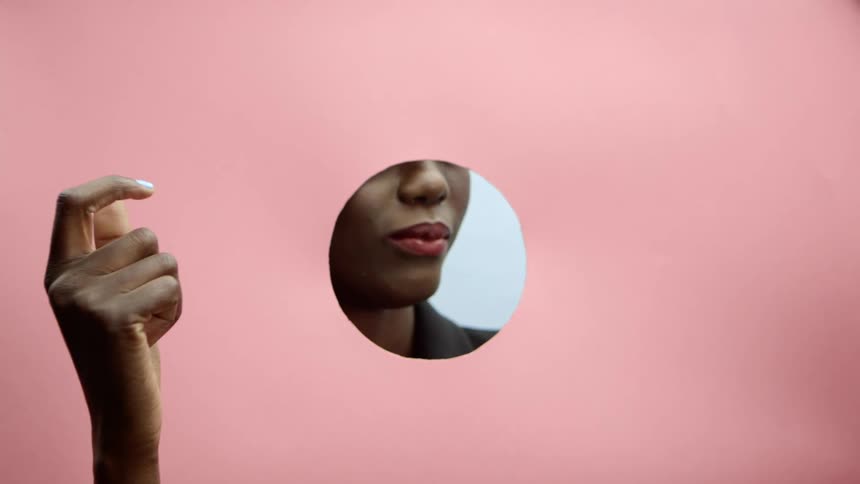 A black womans hand snapping her fingers in front of a pink wall while the lower half of her face can be seen to be on the opposite side of the wall through a hole