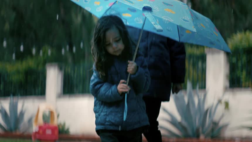 A little girl holding a blue umbrella in the rain while two other girls jump in the rain behind her