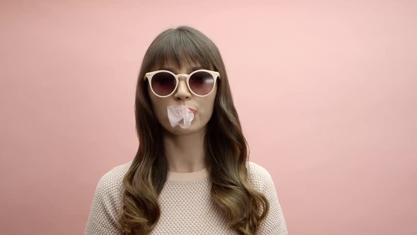 A woman wearing sunglasses and standing against a pink wall chews gum and blows bubbles