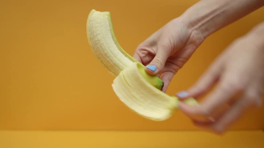 A woman holds a banana and peels the skin back against an orange background