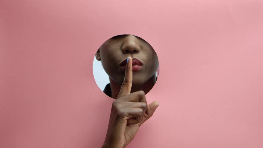 A woman behind a pink paper with a hole hushes with her finger