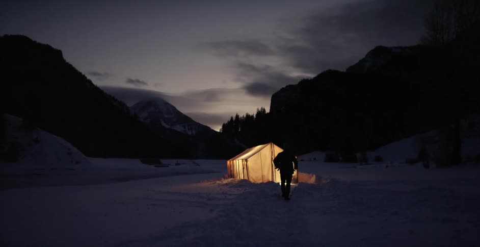 A man walking through snow and entering a brightly lit tent in the mountains