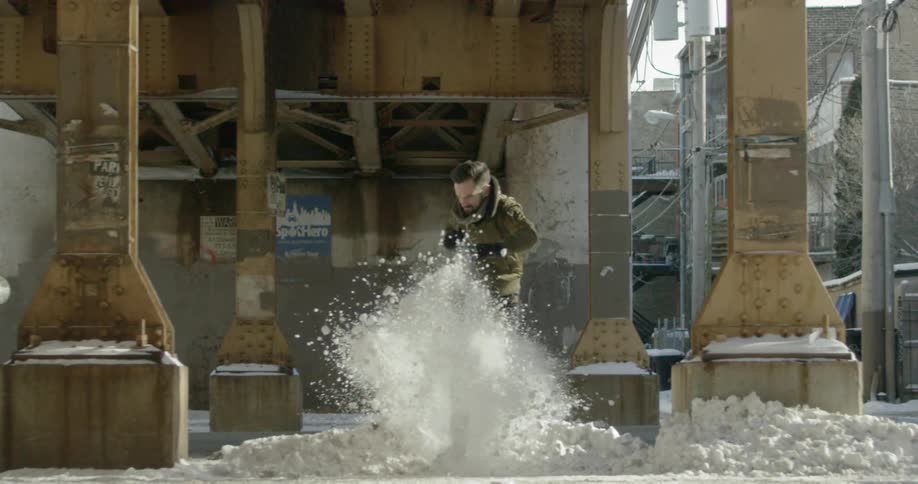 A man runs under a city bridge structure and kicks a pile of snow