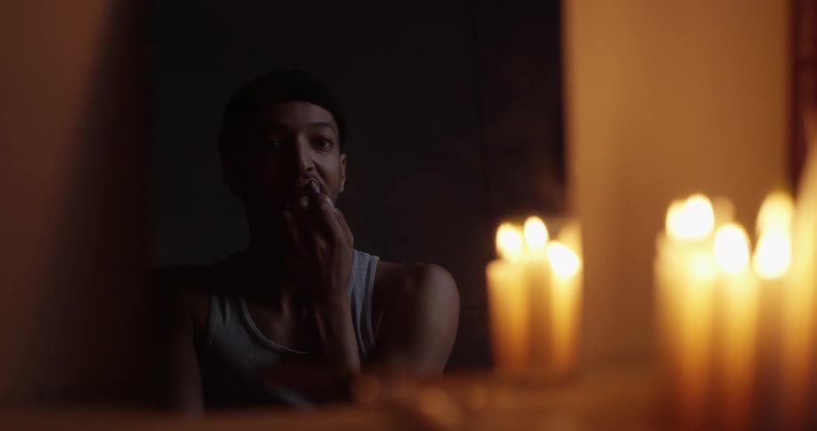 A man applies lipstick while sitting in front of a vanity covered with lit candles