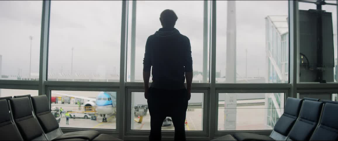 A white male stands at a window in an airport and looks out at the tarmac