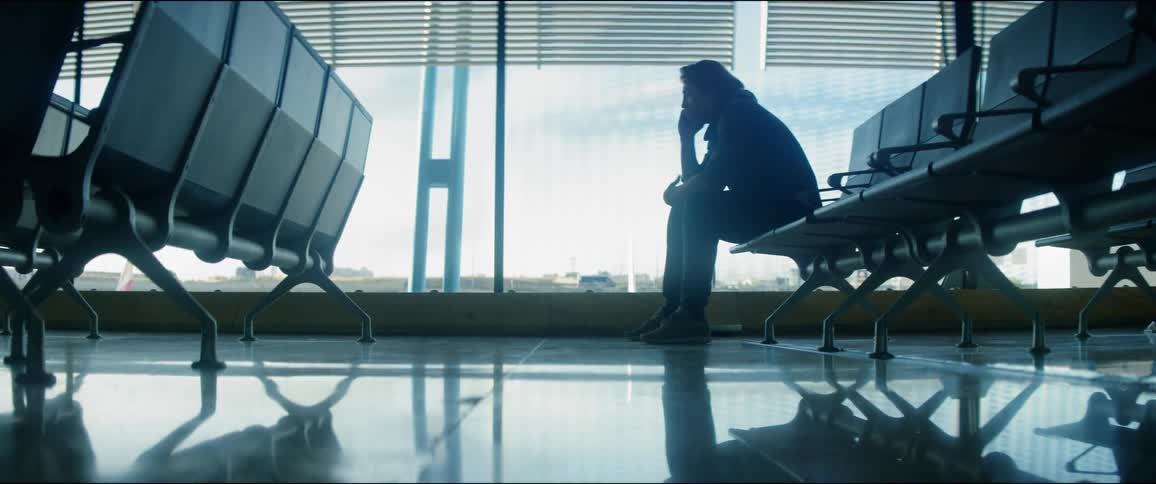 Male sitting alone in a row of aiport seating at the gate next to a window while talking on a cell phone