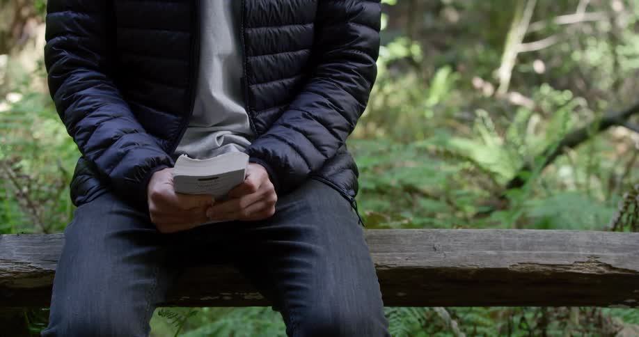 Male sitting on wooden fence rail in the woods removes and book from his back pocket then holds it in front of him and squeezes it tightly