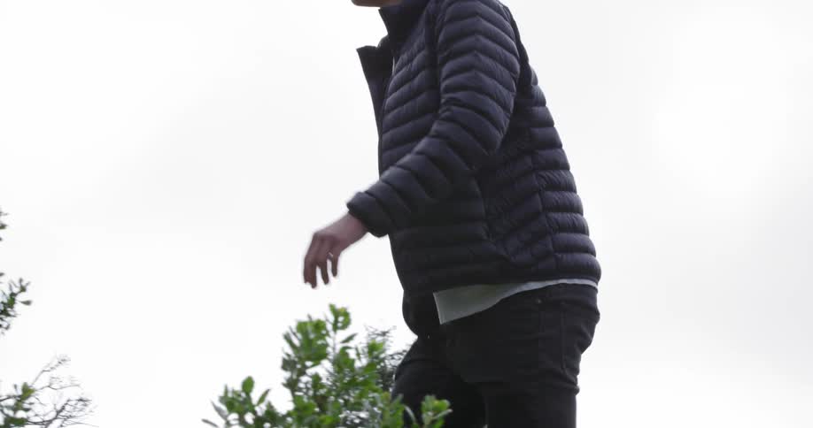 A man looks arounds as he walks up a grassy hill with bushes and trees
