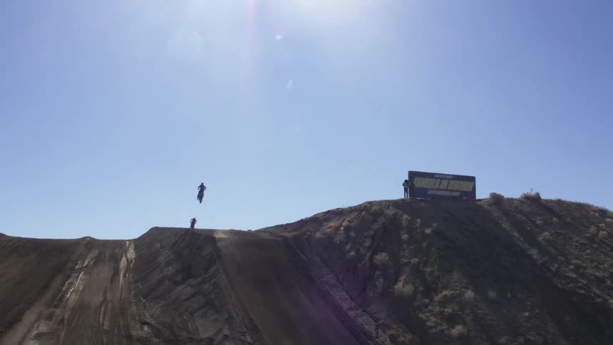 Motorcycles race up and jump off a dirt ramp on a bright blue sky sunny day