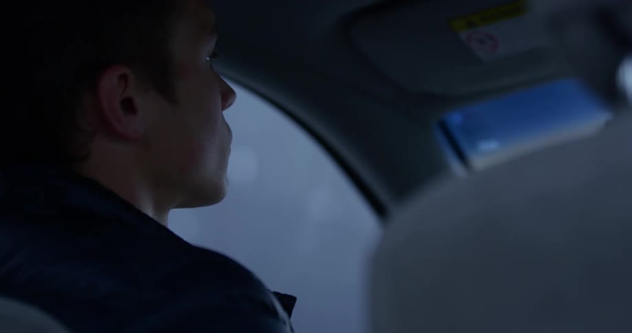 A young man sits in his car and leans his head back and closes his eyes