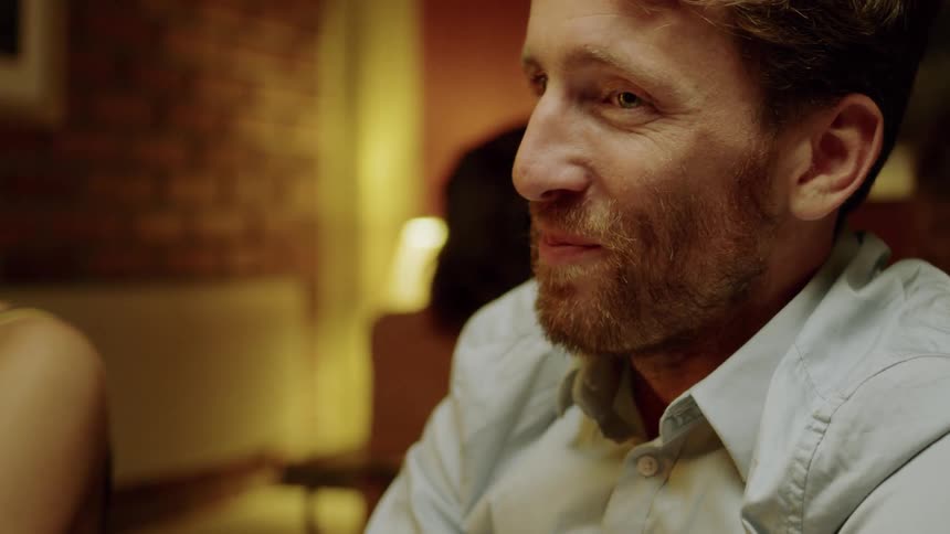 A white male sits with others and breaks into a smile during a conversation