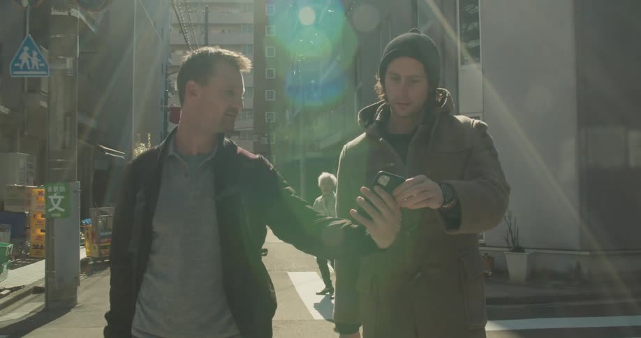 Two men walk along street between buildings studying mobile phone contents